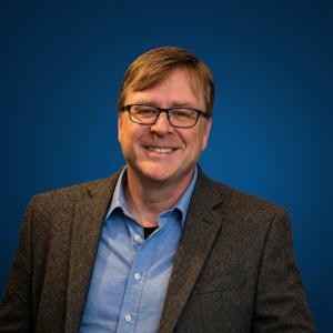 Alt Text: John Wales, Executive Director of the Indiana Township Association, smiles in a professional portrait against a solid blue background. He has short, light brown hair, wears black-framed glasses, and is dressed in a dark textured blazer over a li