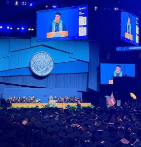 The image is of full arena, with guests flanking three sides and the graduate on the area ground level. It's a sea of students wearing regalia. At the podium is Jim West, speaking about his journey. Behind him is the faculty and leadership of Western Gove