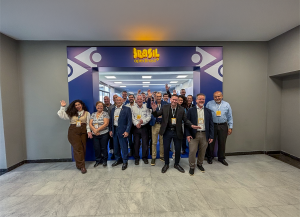 A group of approximately 20 people poses for a photograph indoors, in front of an entrance featuring a blue structure and white graphic elements. At the top, the Brasil logo appears in yellow lettering, with the ApexBrasil logo directly below it. The part