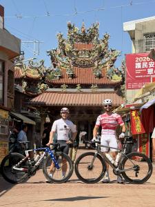 Logan Beck (left) and Gabriel Farmer (right) stand beside their road bikes in cycling gear in front of an ornately decorated traditional Taiwanese temple, with colorful dragon sculptures and intricate roof details.