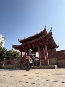 Gabriel Farmer performs a wheelie on his road bike in front of a traditional Taiwanese gate with a red-tiled roof and decorative architectural details under a clear blue sky.