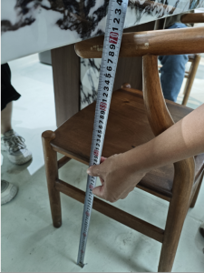 A person holding a tape measure beside a wooden chair beneath a marble-patterned table.