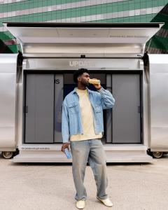 A visitor drinking a can of UPDATE in front of the open Nomad Luxe Walk-In L, its gullwing canopy raised over the sliding doors