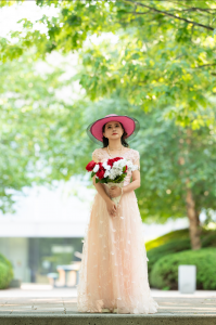 Shekinah Grace Moyes stands beneath leafy trees in a pale pink gown and wide-brimmed pink hat, holding a bouquet of red and white flowers.