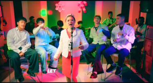  Shekinah Grace Moyes sings at a microphone in a white jacket and red skirt, surrounded by singers and musicians in a studio with colourful lights and Christmas decorations.