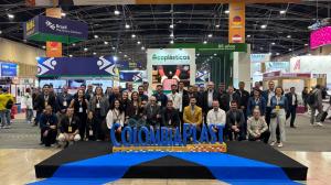 Un grupo de aproximadamente 35 personas posa para una fotografía en el recinto de una feria industrial. Los participantes están distribuidos en dos filas, algunos de pie y otros agachados o sentados, frente a una estructura con el nombre “COLOMBIAPLAST” e