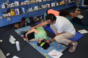 Instructor in white polo shirt and glasses, Aybek Izzatov, kneeling on a blue yoga mat and gently assisting a woman in a green shirt who is lying on her side practicing a muscle-release exercise during a Five Step Sleep Tantra seminar. Two other participa
