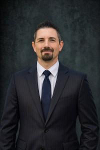 Professional headshot of Eric Carroll, Founder and CEO of Quick Strike Media, wearing a navy pinstripe suit, white shirt and navy tie against a dark textured background.