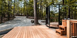 Glass sauna and cedar cold plunge with wooden steps on a deck surrounded by tall pine and hardwood trees.