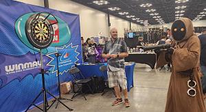 A Winmau dartboard and Championship Darts Corporation display set up at PopCon Indianapolis as a costumed attendee attempts a shot.
