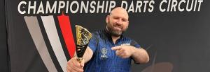 Alex Spellman holds a trophy in front of a Championship Darts Circuit backdrop.