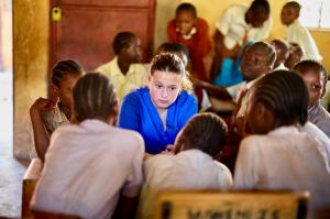 Participant in blue scrubs seated in the middle of a group of schoolchildren gathered around classroom desks.