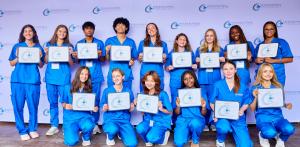 Participants in blue scrubs hold certificates while posing in two rows in front of an International Medical Aid backdrop.