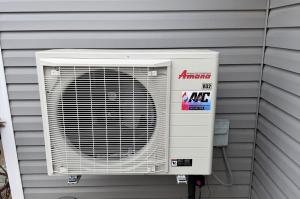 Amana R32 outdoor HVAC unit mounted on brackets against beige vertical siding and a block foundation, with a wooded backyard, installed by All Around Comfort in Cabot, Arkansas.