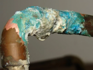 Copper pipe elbow covered in green and white corrosion buildup with a water drip forming