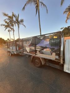 A white rubbish removal truck with a large metal cage trailer attached, both loaded with bulky household junk and unwanted items for disposal. The truck and trailer are filled with discarded furniture, including sofas, cushions, mattresses or padded furni