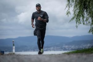 A runner in dark athletic clothing running along a waterfront path with trees and an overcast sky in the background.