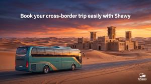Bus on a desert highway at sunset passing a traditional mud-brick fort with wind towers, with text "Book your cross-border trip easily with Shawy"