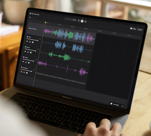 A laptop on a wooden café table shows the Nomono Studio Cloud multitrack editor. Three color-coded speaker tracks, Blue, Green and Purple, sit above a gray Ambience track. Each track has its own volume slider.