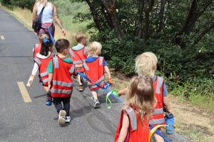 Line of young children in red safety vests walking along a paved path with a teacher