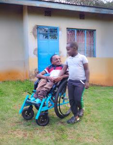 An African man, seated in a blue wheelchair, looks lovingly at his 8-year-old son, who stands proudly next to his wheelchair, in Kenya.