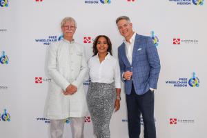 A man in a white suit, a woman in a white blouse and sparkling skirt, and a man in a tailored blue suit pose in front of a step-and-repeat at a fundraising gala.