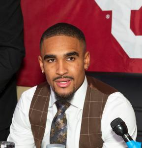 Jalen Hurts seated at a press event, wearing a white shirt, brown plaid vest, and patterned tie, with a microphone positioned in front of him.