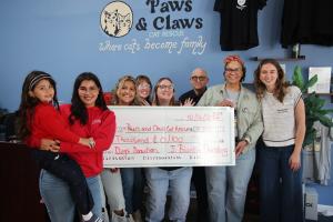 Nine people stand in front of a blue wall painted with the Paws & Claws Cat Rescue logo and the words “Where cats become family.” They hold an oversized check dated 10/16/2026, made out to Paws and Claws Cat Rescue for $5,000 from J. Blanton Plumbing. On 