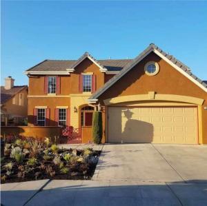 Two-story residential home with warm terracotta and beige stucco exterior, dark shutters, concrete driveway, and front yard landscaping under a clear blue sky.