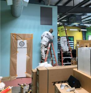 Two professional painters on ladders applying light blue paint to a commercial wall in a retail space with masked doorways and equipment boxes.
