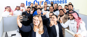 Diverse group of colleagues smiling and giving thumbs-up while posing for a selfie in an office.