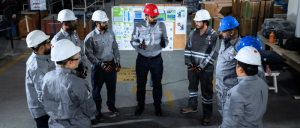 Factory workers in hard hats gather around a supervisor for a safety briefing in an industrial warehouse.