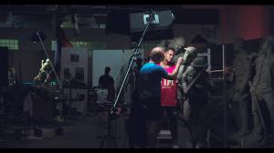 Sabin Howard works on a clay soldier’s face beside a live model in a red shirt, under studio lights.