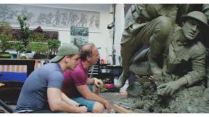 Two men sit beside clay soldiers in dynamic battle poses, studying the sculpted figures at close range.