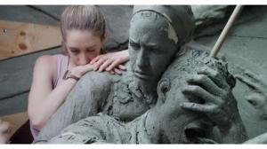 A woman rests her hands and face against the shoulder of a clay soldier holding his helmet.