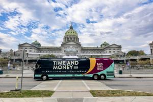 A large bus in bright colors and with the text "Time and Money Tour" parked in front of the Pennsylvania State Capitol.