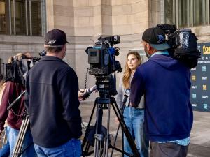 A woman in a gray jacket and jeans stands in front of cameras.