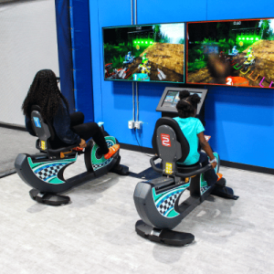 Two children play interactive racing games on colorful simulator machines in a brightly lit arcade.