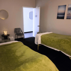 A cozy twin-bed room with green blankets, an open door, black chairs, candlelit table, and framed landscape art.
