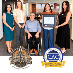 Five people pose indoors, with a seated man in a wheelchair and a framed award, alongside Beyond Words Bookshop and Certified Autism Center logos.