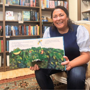 Smiling woman sits in a bookstore, holding open a colorful illustrated children’s book featuring a green monster landscape.