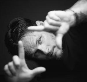 Black-and-white actor headshot of a man with swept-back hair and facial hair, using his hands to frame his face against a dark studio background, creating a dramatic, artistic, and expressive portrait.