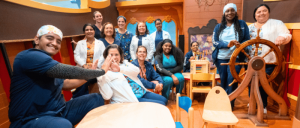 Group of smiling people posing in a colorful ship-themed room, with one person seated at a large wooden ship’s wheel.