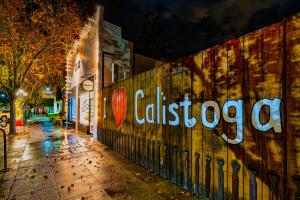 A large wooden fence painted with “I Heart Calistoga” along a rain-soaked sidewalk in downtown Calistoga at night, illuminated by warm storefront lights and autumn-colored trees.