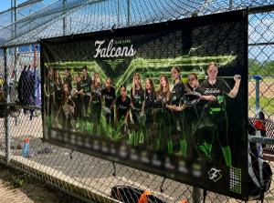 Large black Oak Park Falcons softball banner featuring players in black and green uniforms, displayed on a chain-link fence at a softball field.