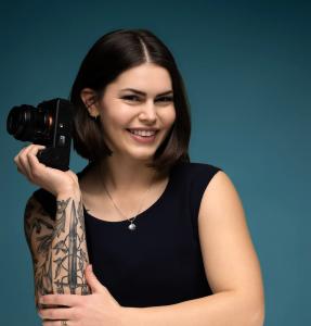 Professional portrait of a smiling female photographer with a dark bob haircut and tattooed arm, holding a camera against a teal studio background.