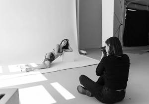 Black-and-white behind-the-scenes photo of a photographer capturing a boudoir model posing on a studio backdrop, with natural light streaming across the floor.