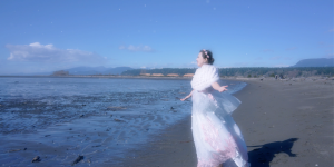 Shekinah Grace Moyes stands at the shoreline with her arms open, wearing a flowing white and pink gown, with ocean water and distant mountains behind her.