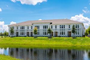 A row of two-story townhomes with light gray and white siding, a shingled roof and wood front doors sits behind a green lawn lined with young trees. A pond in the foreground reflects the buildings under a blue sky with scattered clouds.