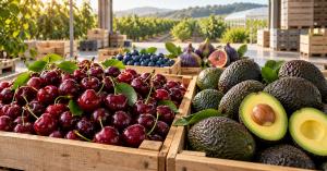 Cherries and avocados in wooden crates, with blueberries and figs behind them, overlooking an orchard and greenhouse.